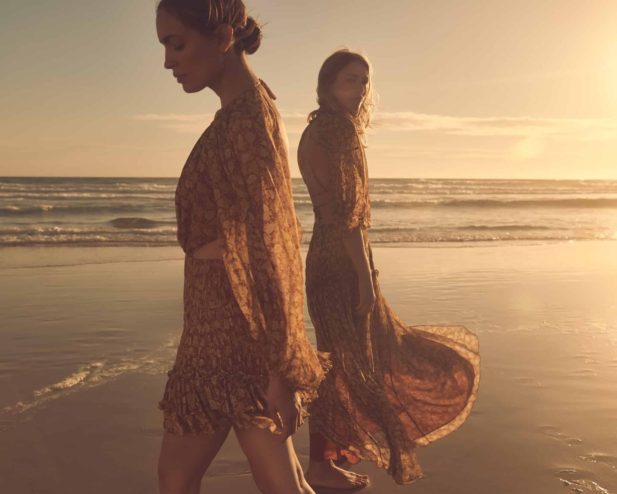 Shona Joy campaign image - two female models wearing floral print Shona Joy dresses, walking in opposite directions on the wet sand ofn a beach at low tide, sunset.