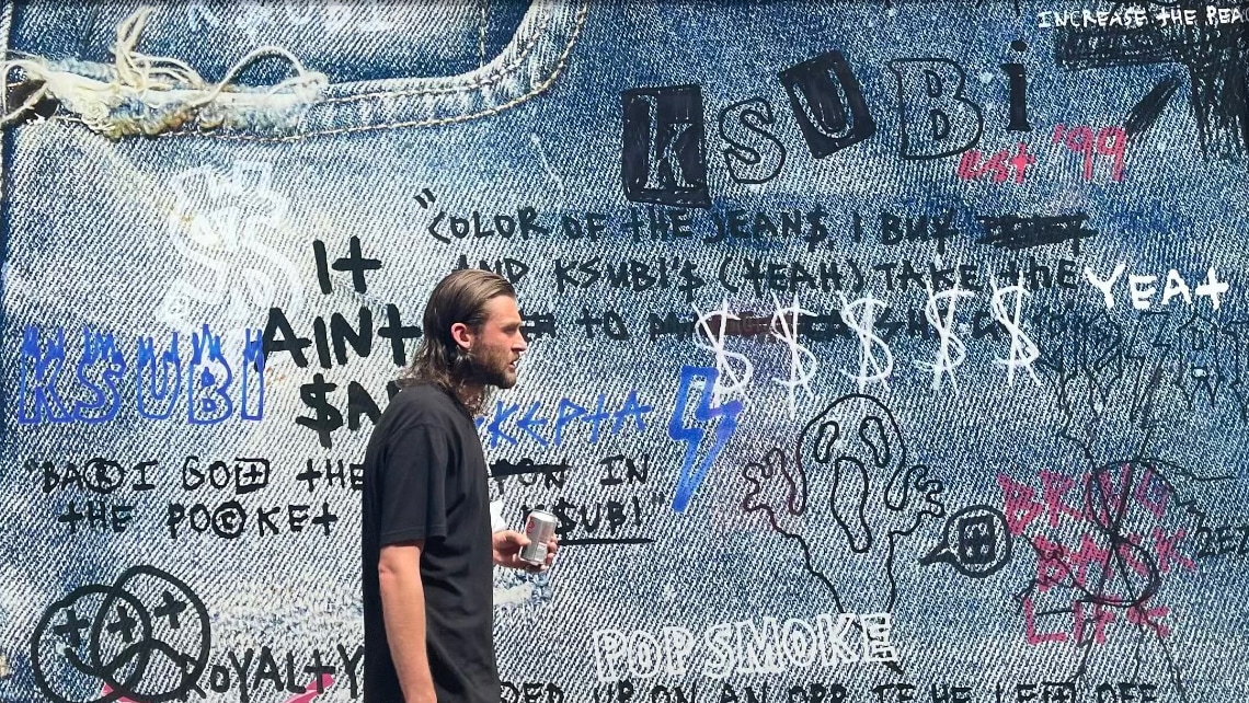 A young man with long brown hair and beard wearing a black t-shirt walking past the window hoarding of the under-construction Ksubi store in Carnaby St, London. The hoarding features a close-up image of Ksubi jeans covered in Ksubi brand symbols and handwritten lyrics from songs that mention Ksubi.