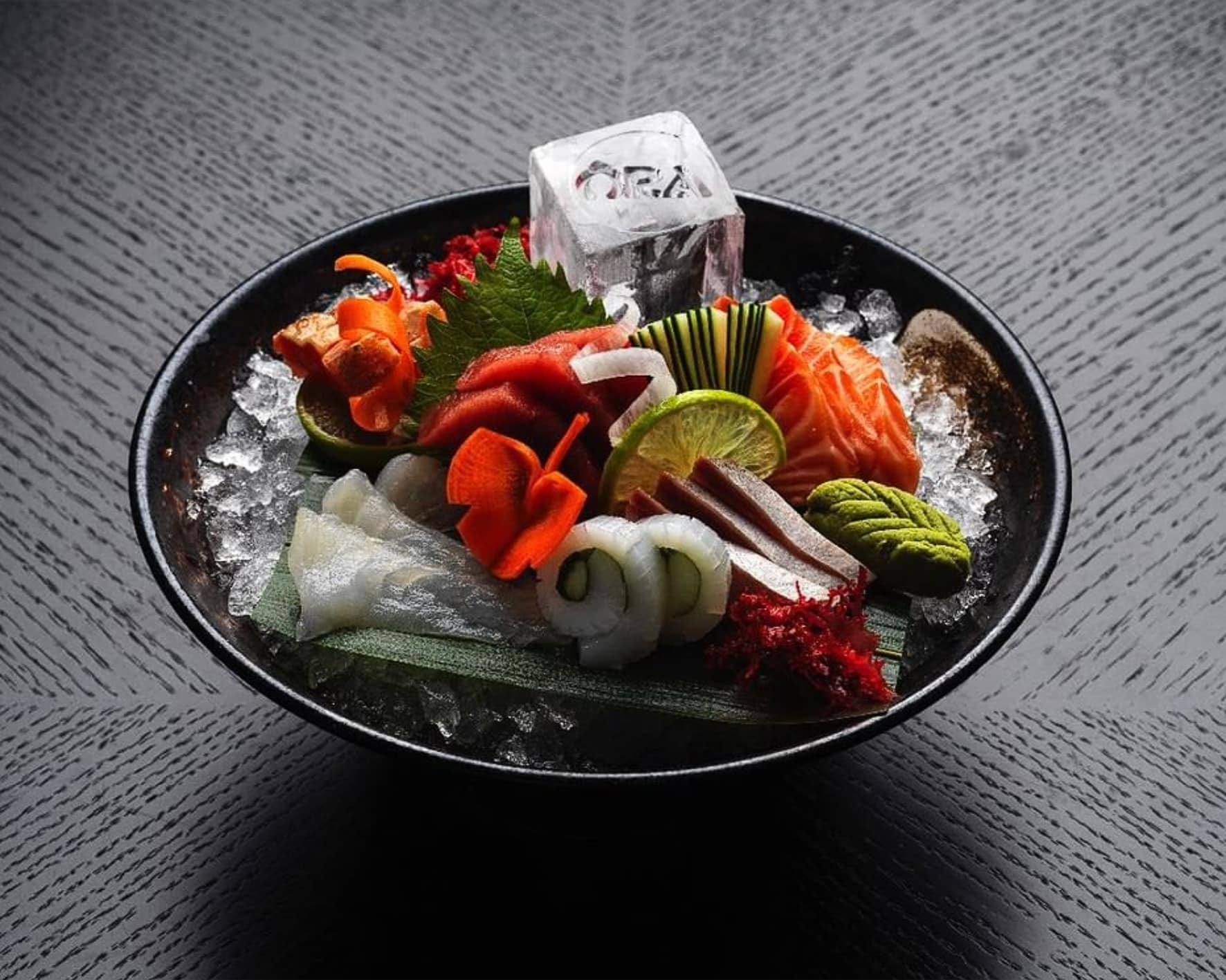A black bowl sitting on a black wood surface, filled with a variety of glistening fresh sushi and sashimi. A large ice cube with the Ōra logo engraved into it is nestled amongst the sushi and sashimi.
