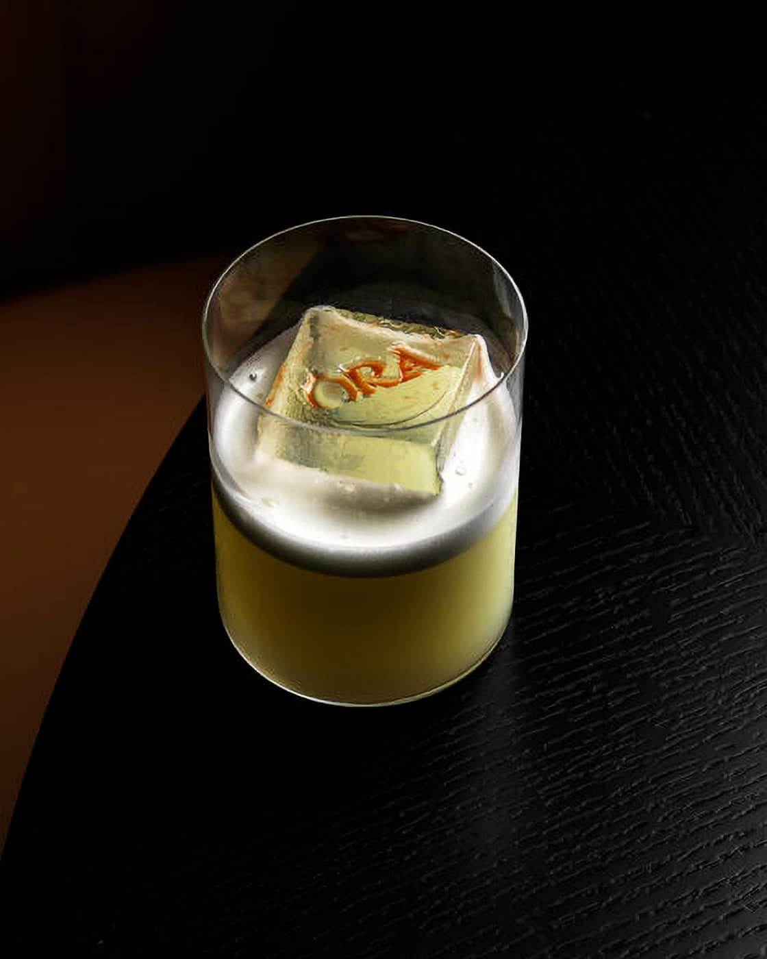 Overhead shot of a cocktail in a tumbler glass sitting on a black wooden table. The cocktail has a white foam on top of the liquid, and a large ice cube with the Ōra logo engraved into it floats in the glass.