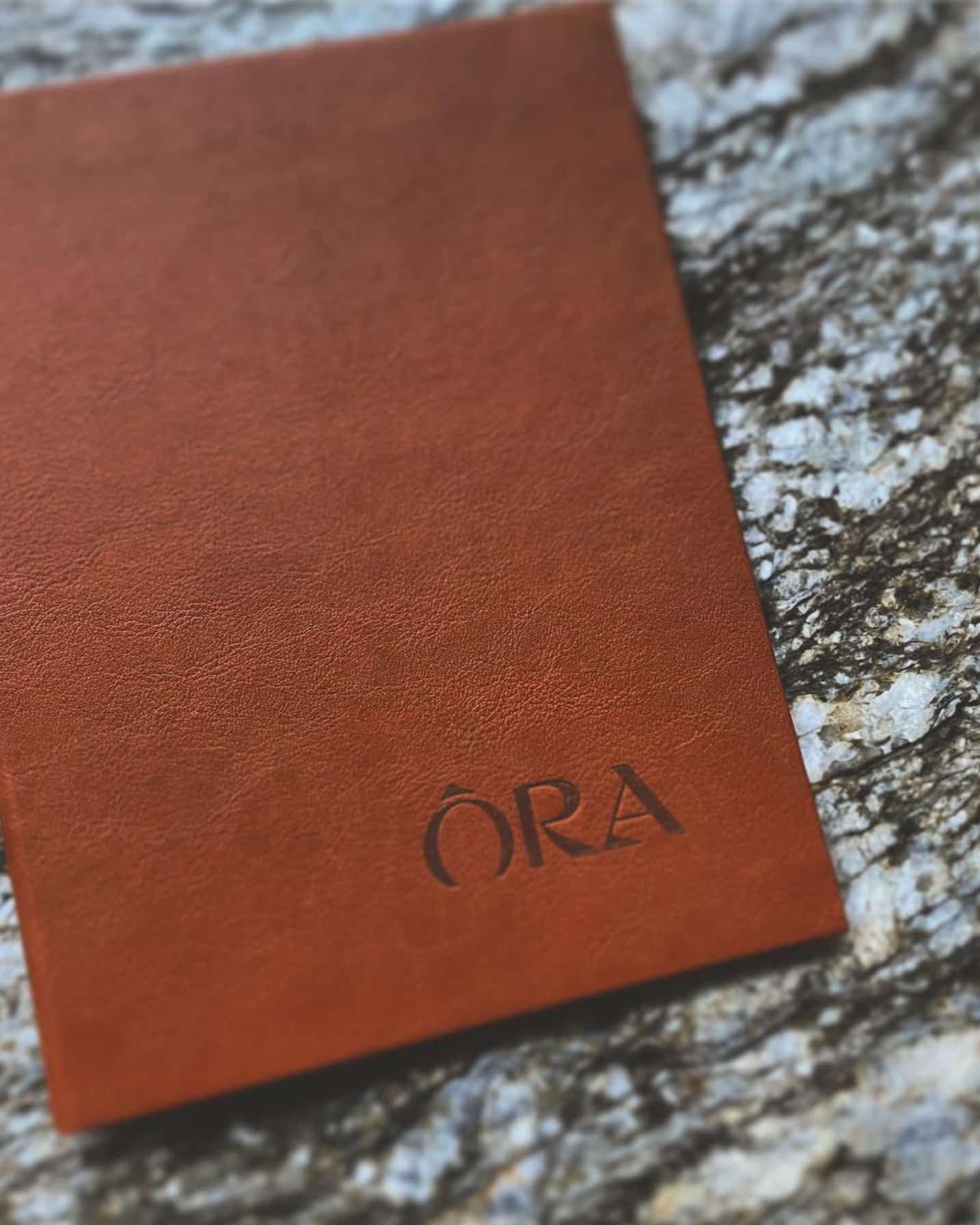 A brown leather menu cover, bearing the Ōra logo, sits on a mottled granite surface.