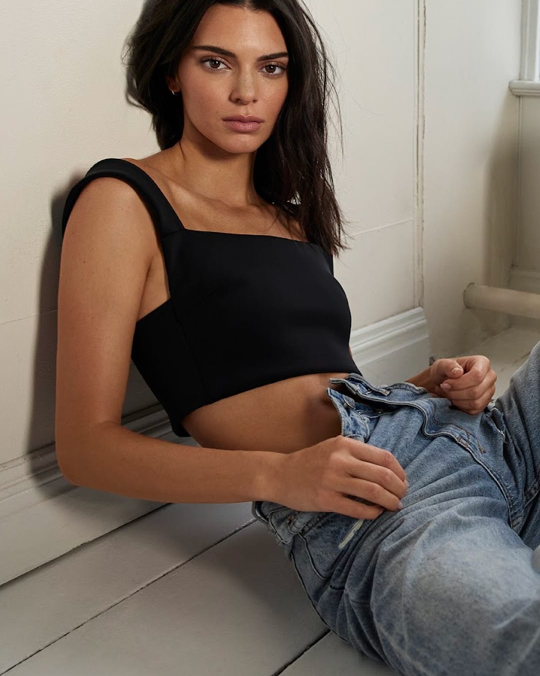 Kendall Jenner wearing a black crop top and mid-blue washed Ksubi jeans sitting on the floor of a white room, leaning back on the wall. The floor is white painted floorboards. A white radiator with peeling paint and a window with a white gauze curtain is in the background.