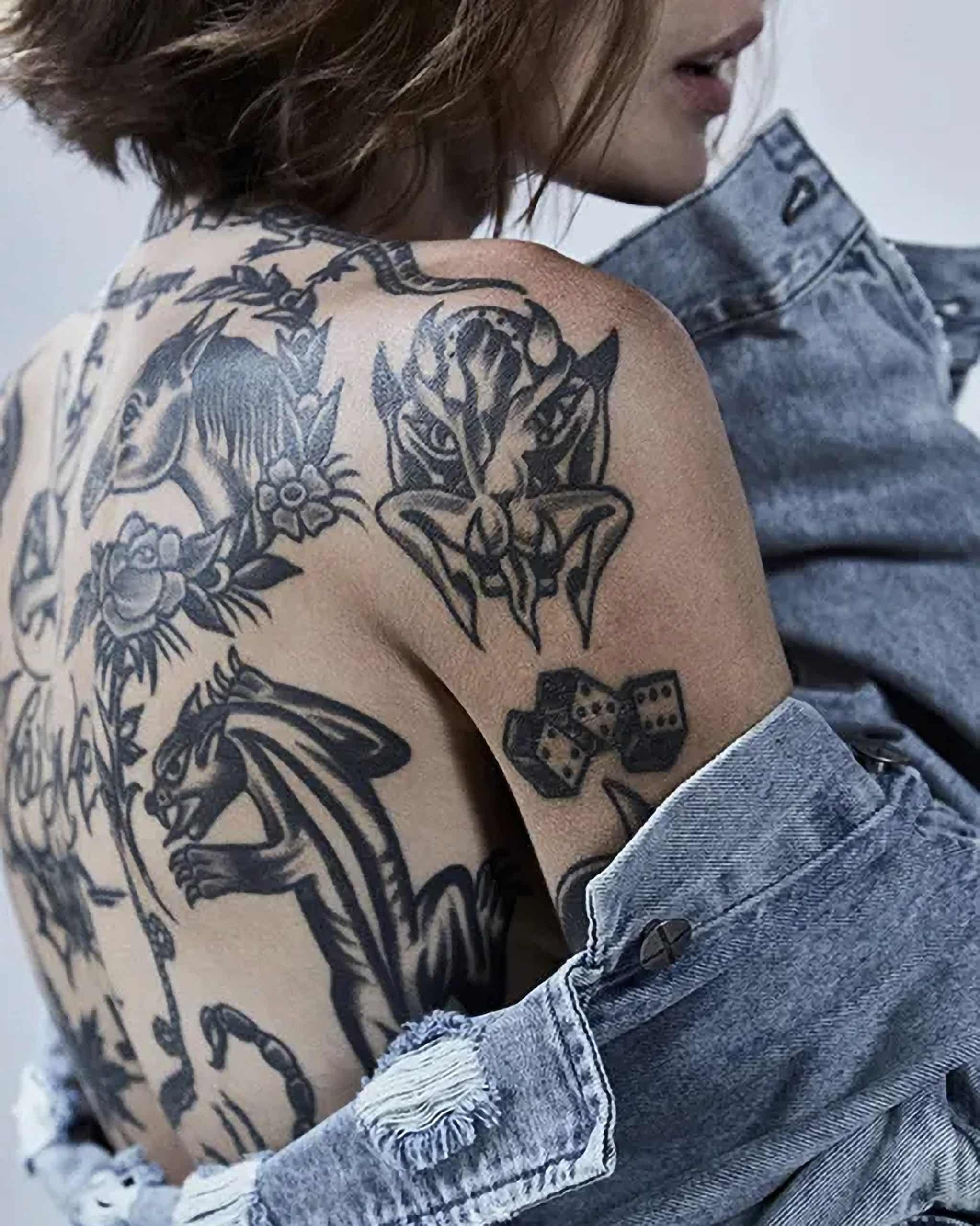 A photograph of a female model with brown shoulder-length hair, wearing a light blue Ksubi denim jacket which is falling off her shoulders. The model's shoulders and upper back is visible and are covered in classic-style tattoos.