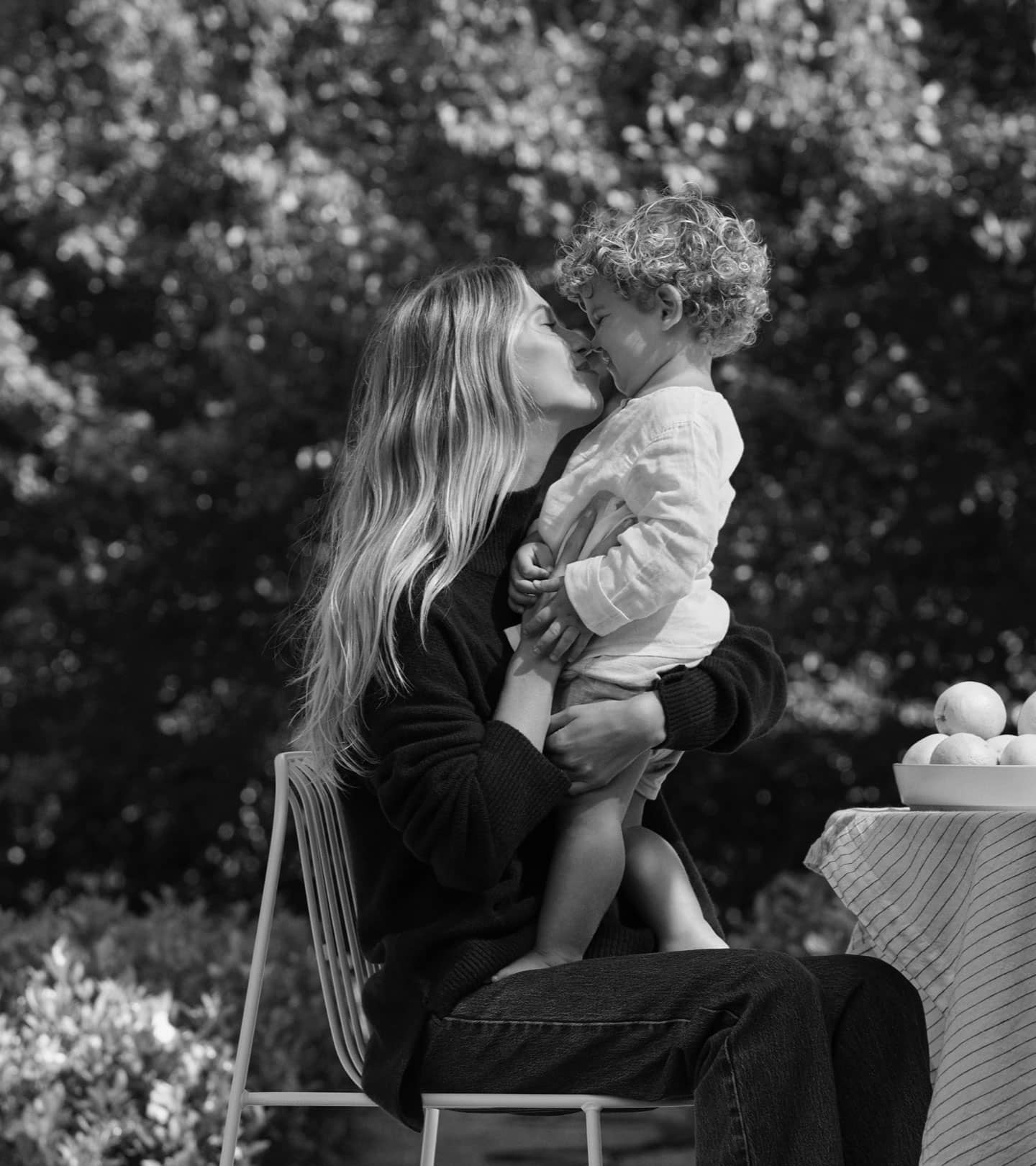 A mother and her toddler, sitting on a chair in a garden, next to a table. The toddler is standing on their mother's lap, and they are touching noses.