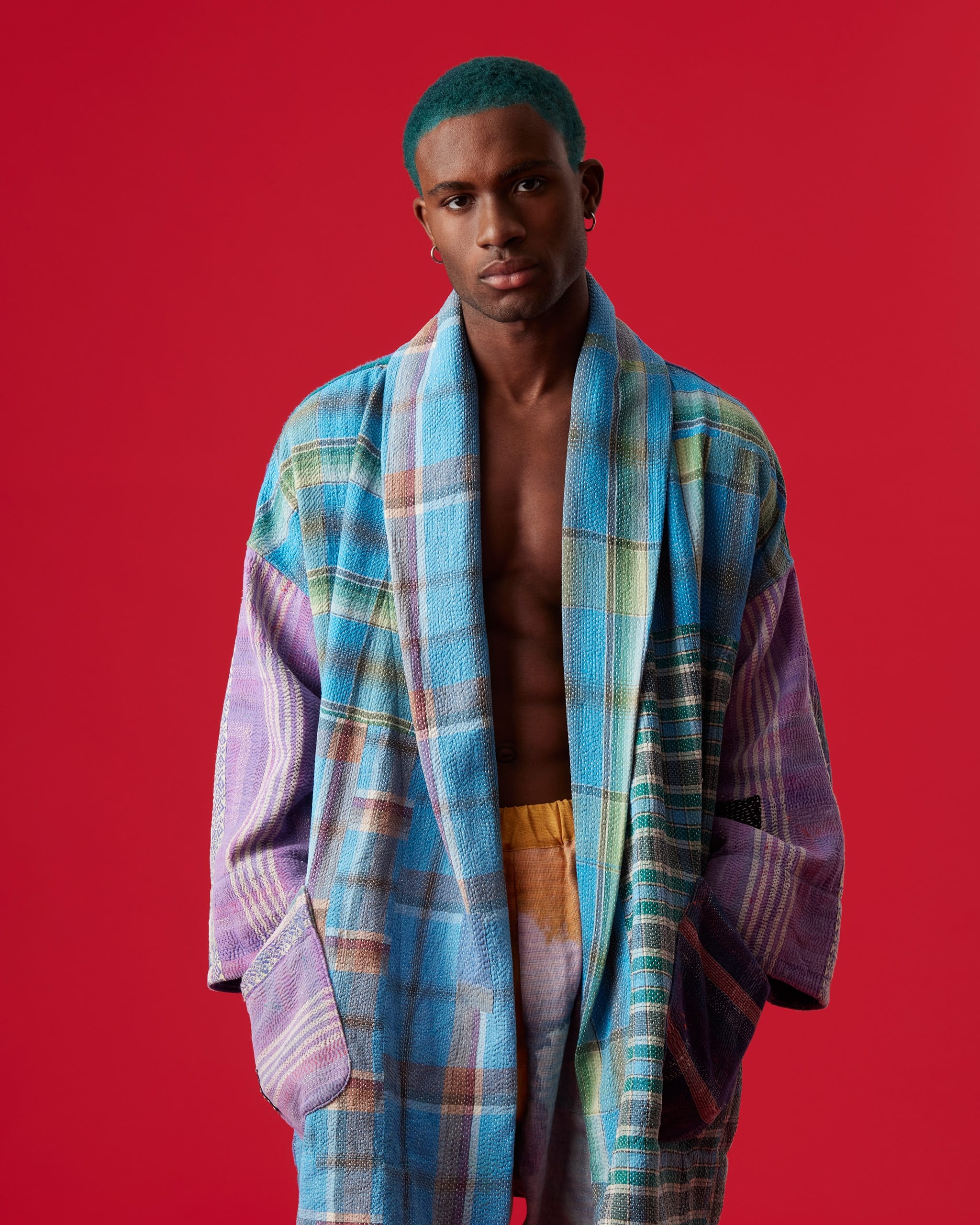 A male model with close-cropped hair that has been dyed blue looks directly at the camera, wearing a multi-coloured plaid overcoat. His hands are in the overcoat pockets, and he is standing in front of a solid red wall.