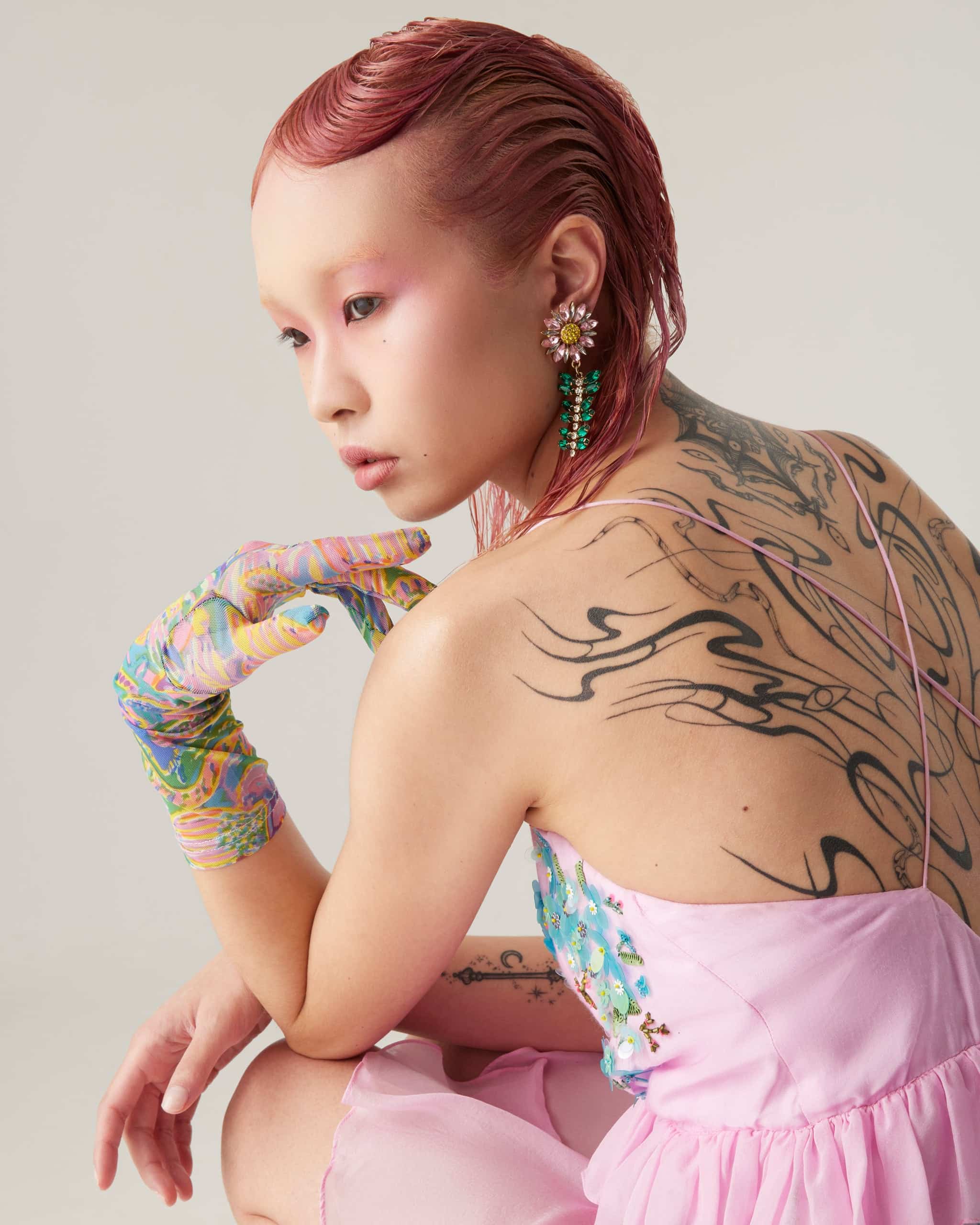A female model with slicked-back mid-length pink hair crouching down, wearing a pink Romance was Born dress and multi-coloured gloves. She has a large tattoo of wavy black lines covering her back.