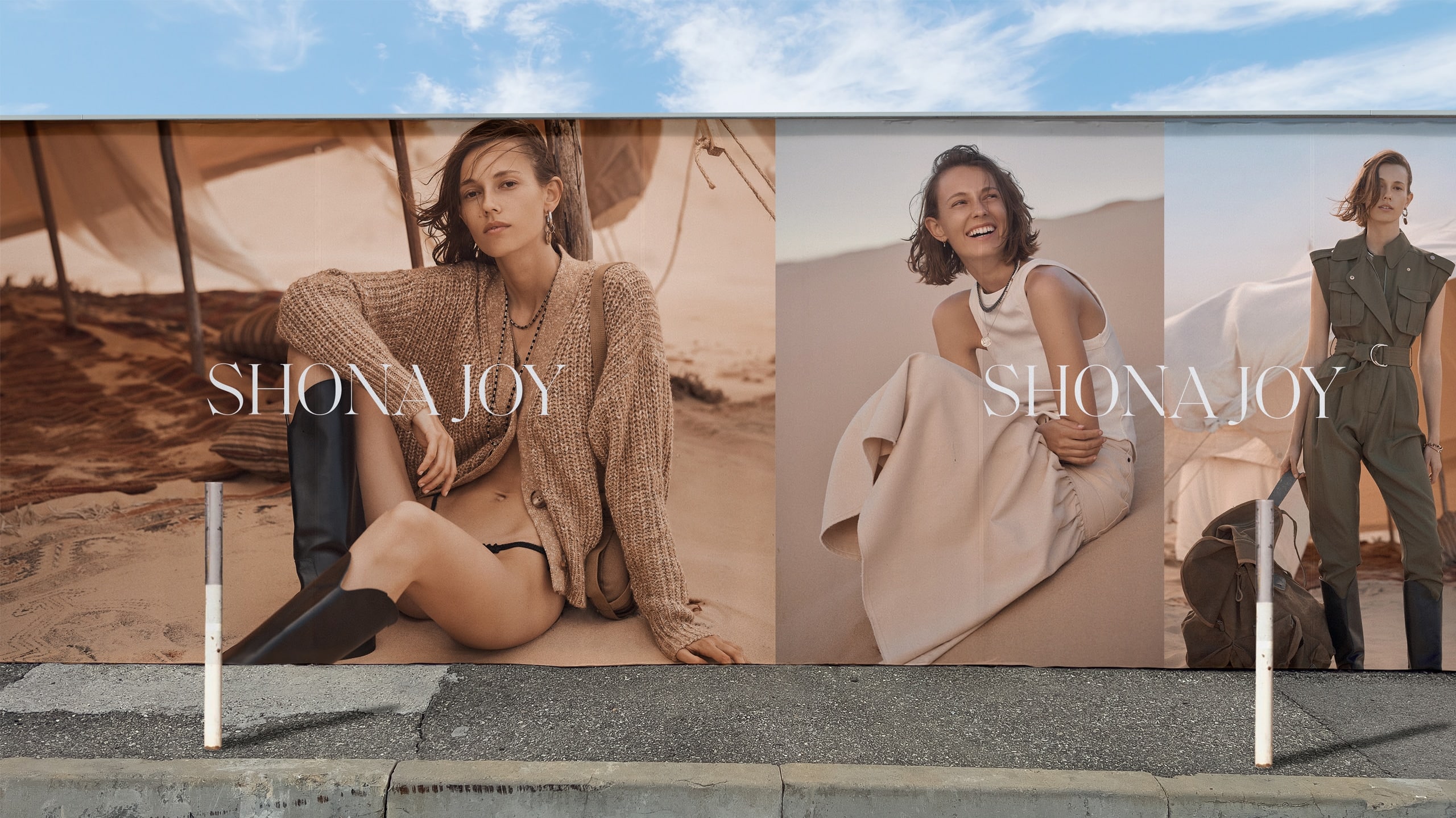 Posters on a wall showing a Shona Joy campaign. On the posters are three different photographs of a female model sitting and standing on sand dunes with a large canvas tent that is billowing in the wind in the background.
